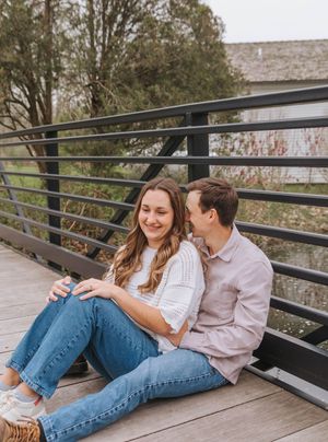 Couple sitting on a wooden bridge, woman in white top and jeans laughing as man in pink shirt whispers to her.