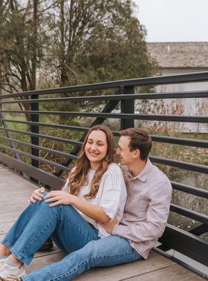 A smiling couple sits together on a wooden bridge, sharing a joyful moment with lush green trees in the background.