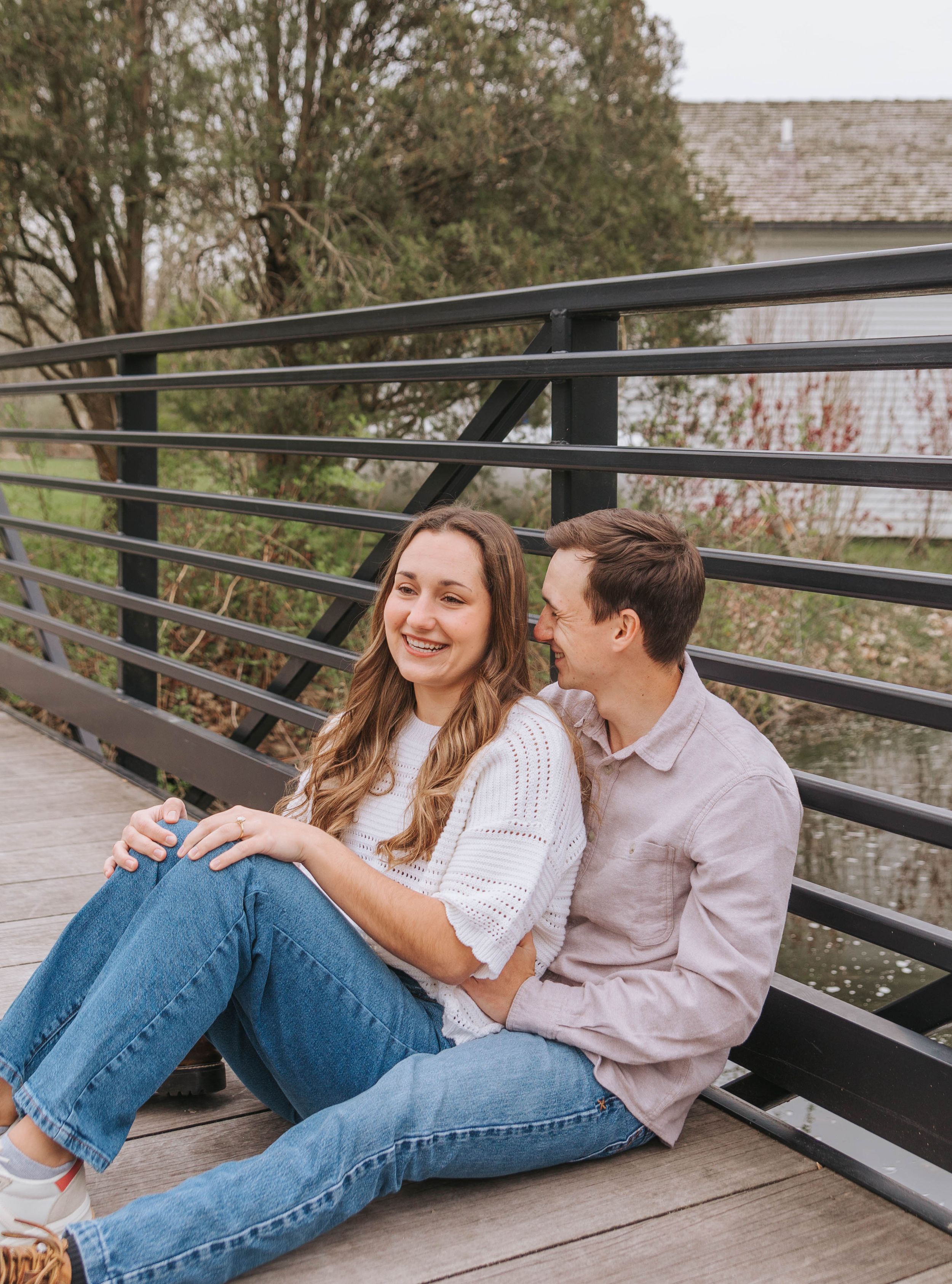 Happy couple sitting together on a wooden bridge, woman smiling in white top and jeans, man in a pink shirt.