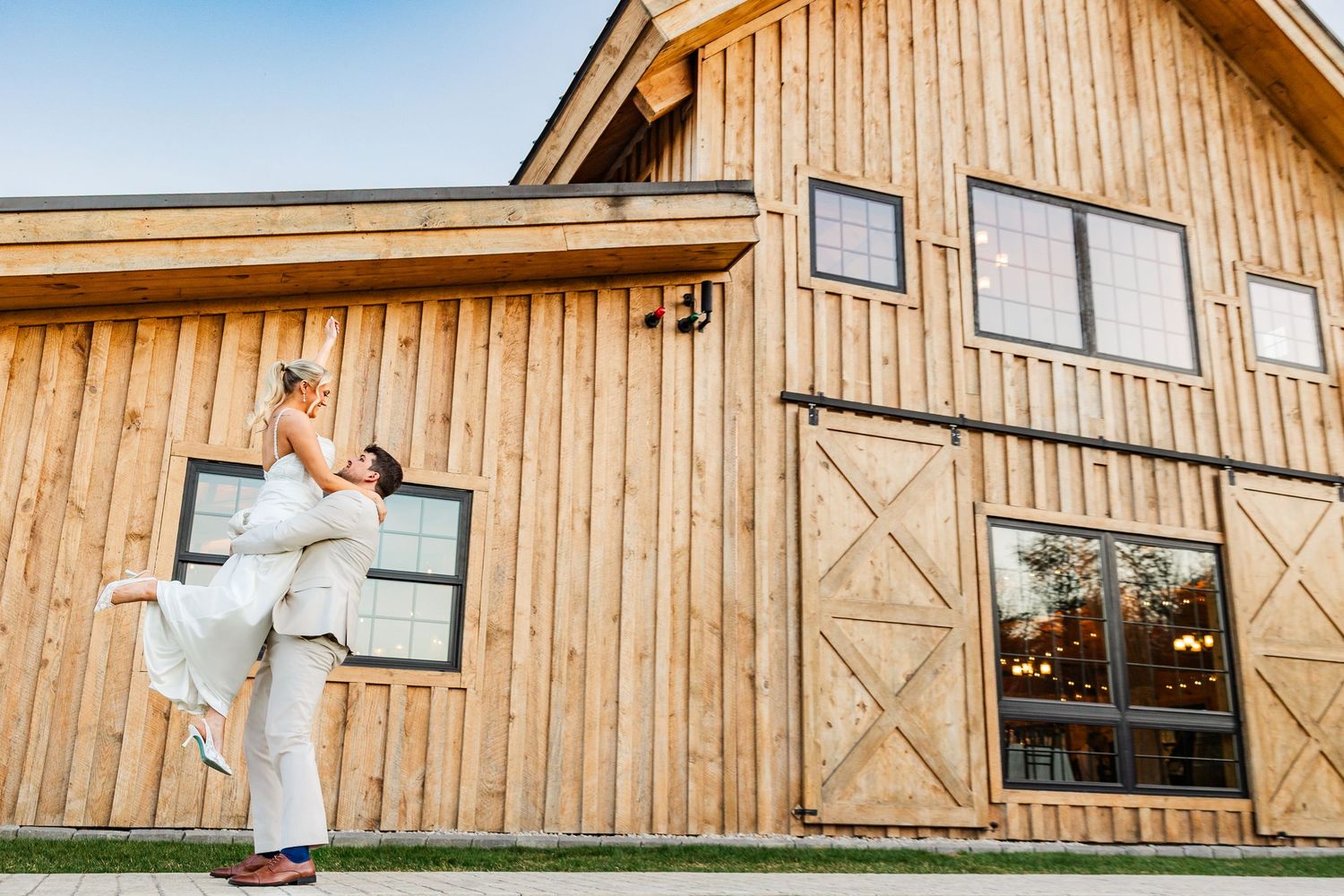 Groom lifting bride in white dress outside rustic wooden barn wedding venue at golden hour.