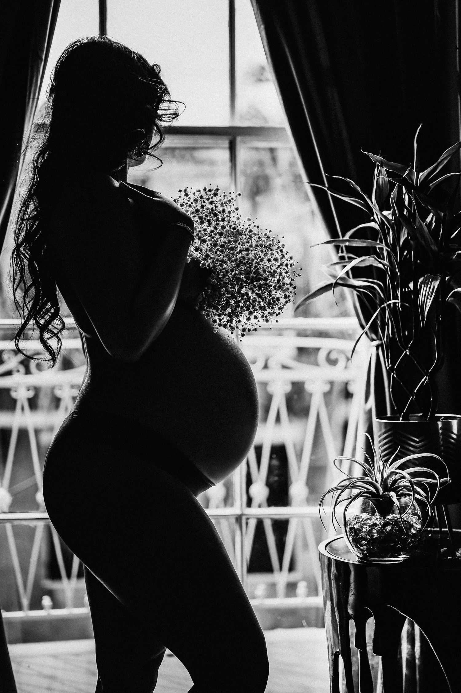 Silhouette of pregnant woman holding flowers standing by window, artistic black and white maternity photo. pregnancy boudoir photoshoot