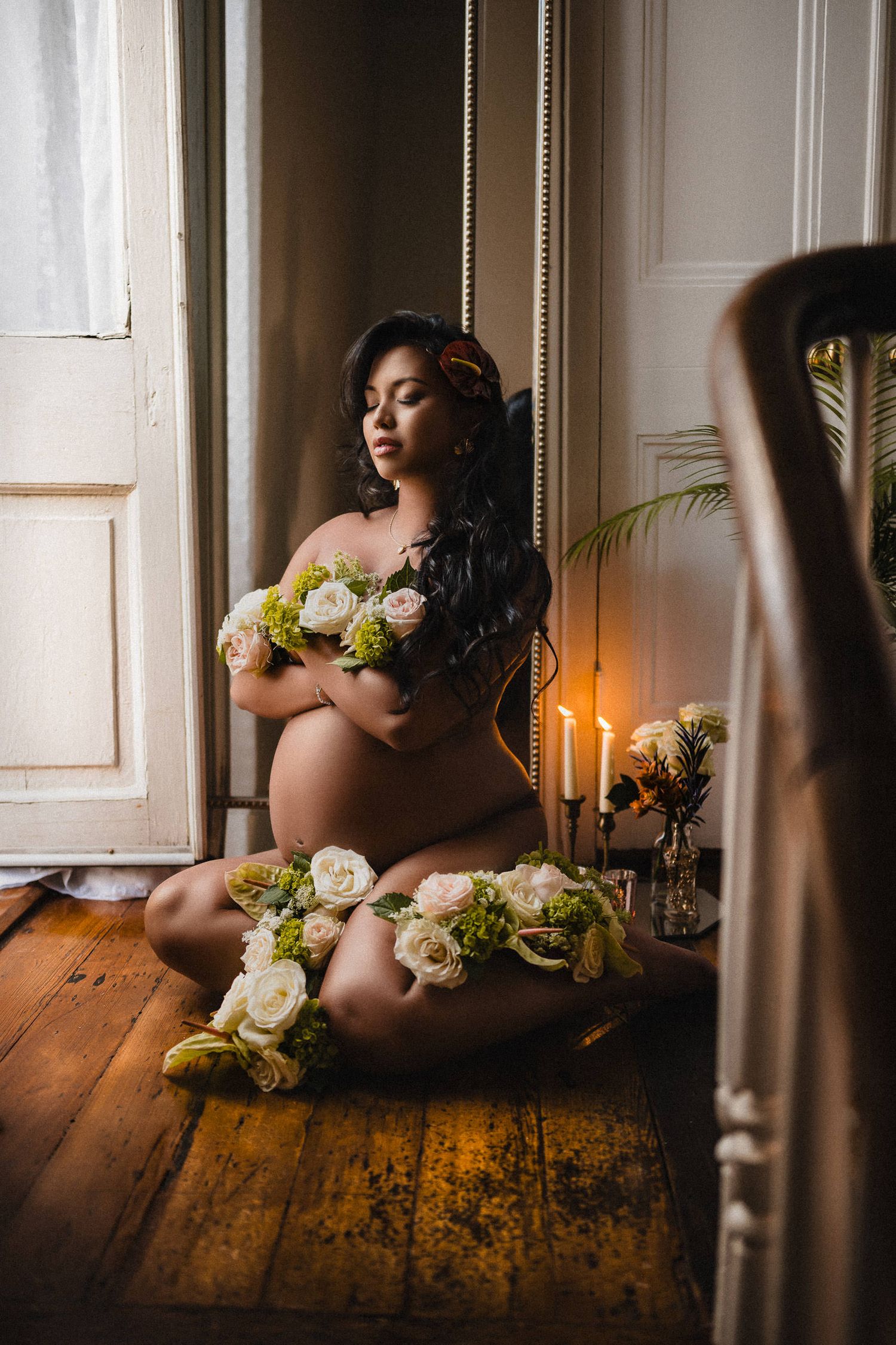 Artistic maternity photo featuring white roses and candlelight in a moody setting with wooden floors. luxury maternity boudoir photos.