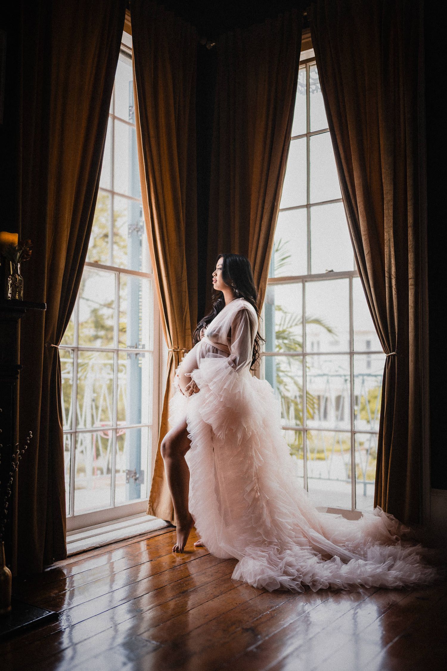 Maternity portrait of woman in flowing pink tulle gown standing by tall windows with brown drapes. maternity boudoir session ideas.