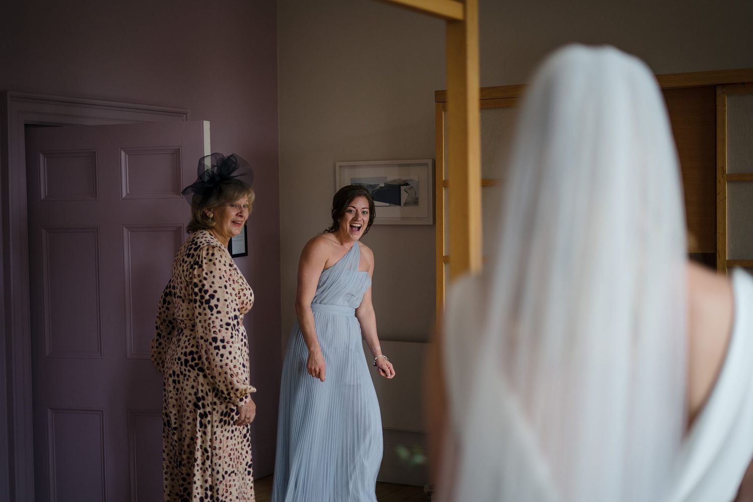 Bride reveals wedding dress to emotional bridesmaids in light blue gowns during bridal preparation.