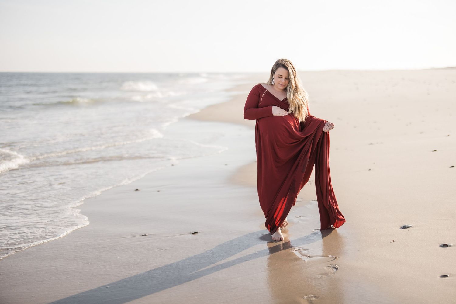 Woman in red dress posing on sandy beach with ocean waves, barefoot in shallow water on a sunny day.