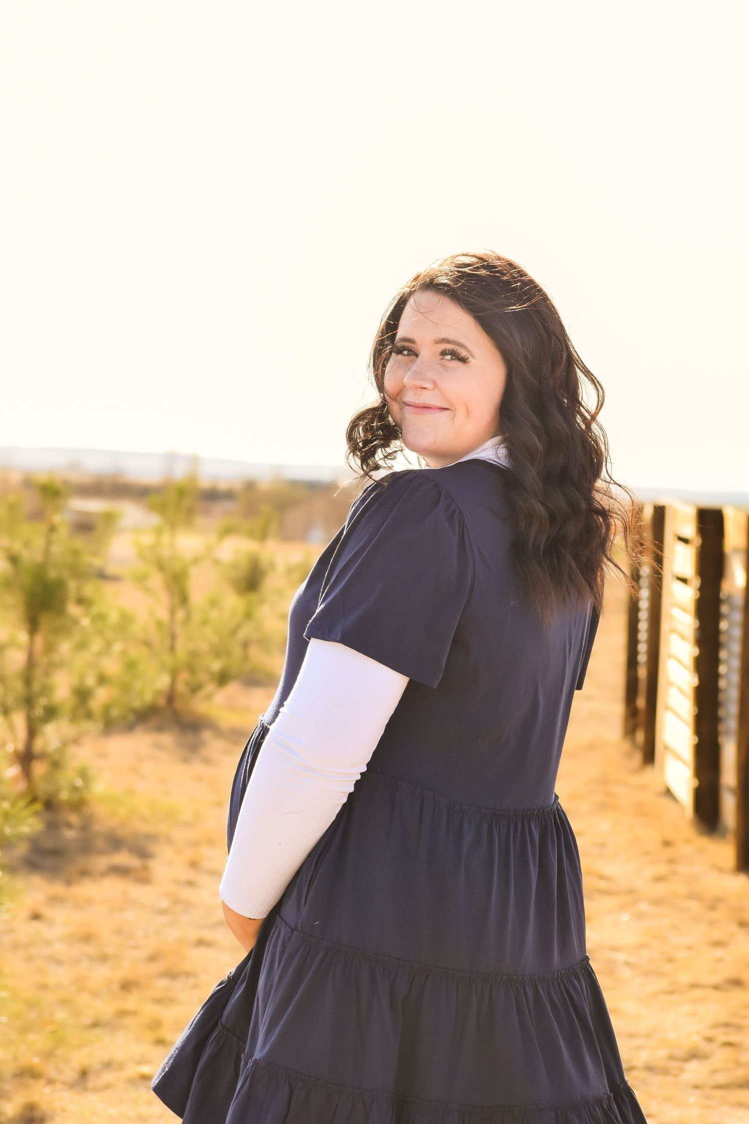 A woman in a navy tiered dress with white sleeves smiles over her shoulder outdoors near a fence.