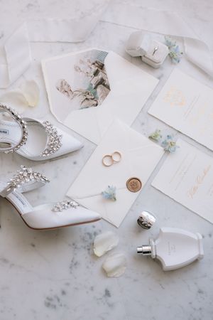 Elegant wedding flat lay with crystal heels, gold rings, wax seal invitations, perfume bottles, and delicate blue flowers on marble.