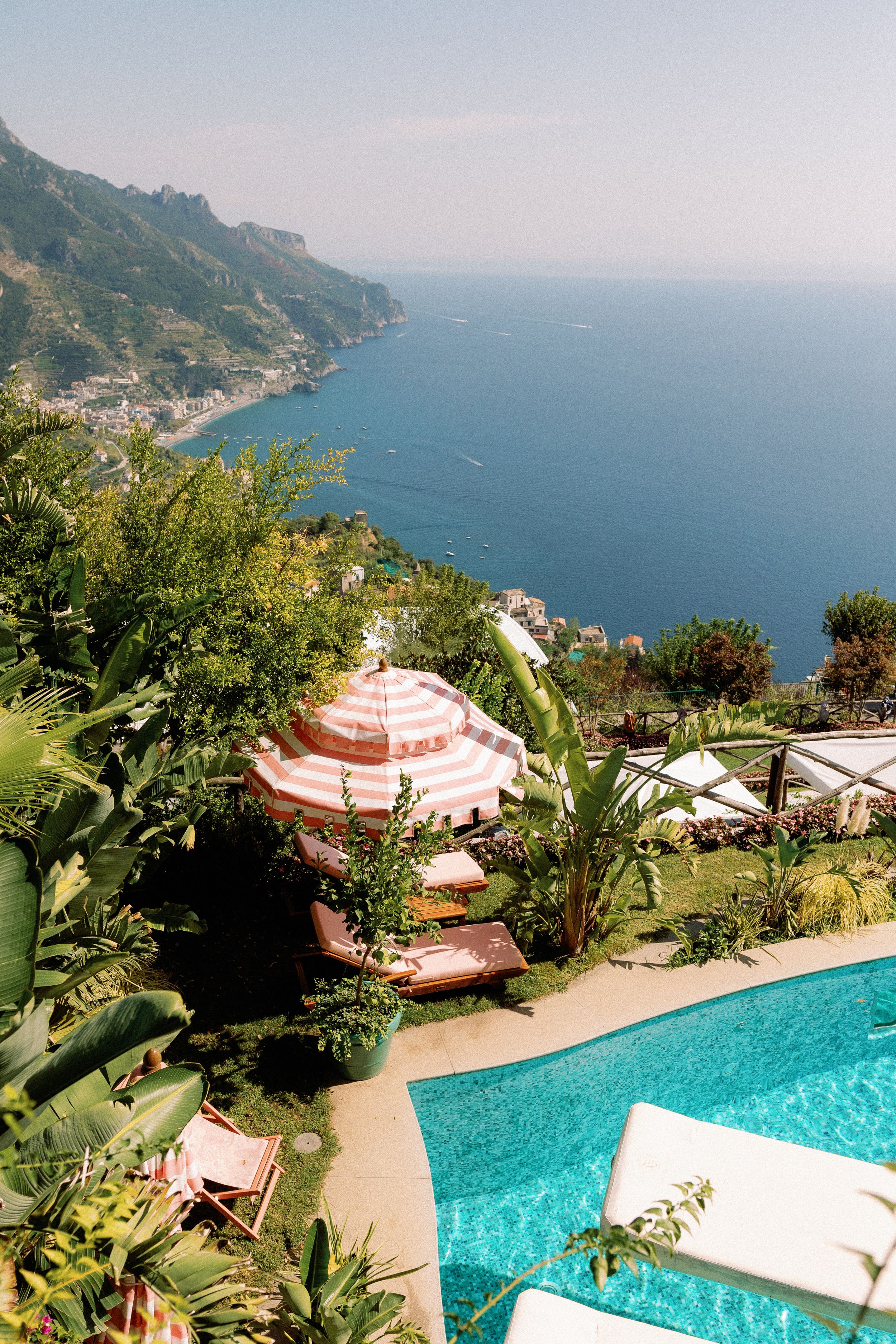 Luxury pool terrace with striped umbrellas overlooking lush gardens and stunning Amalfi Coast sea views on a sunny day.