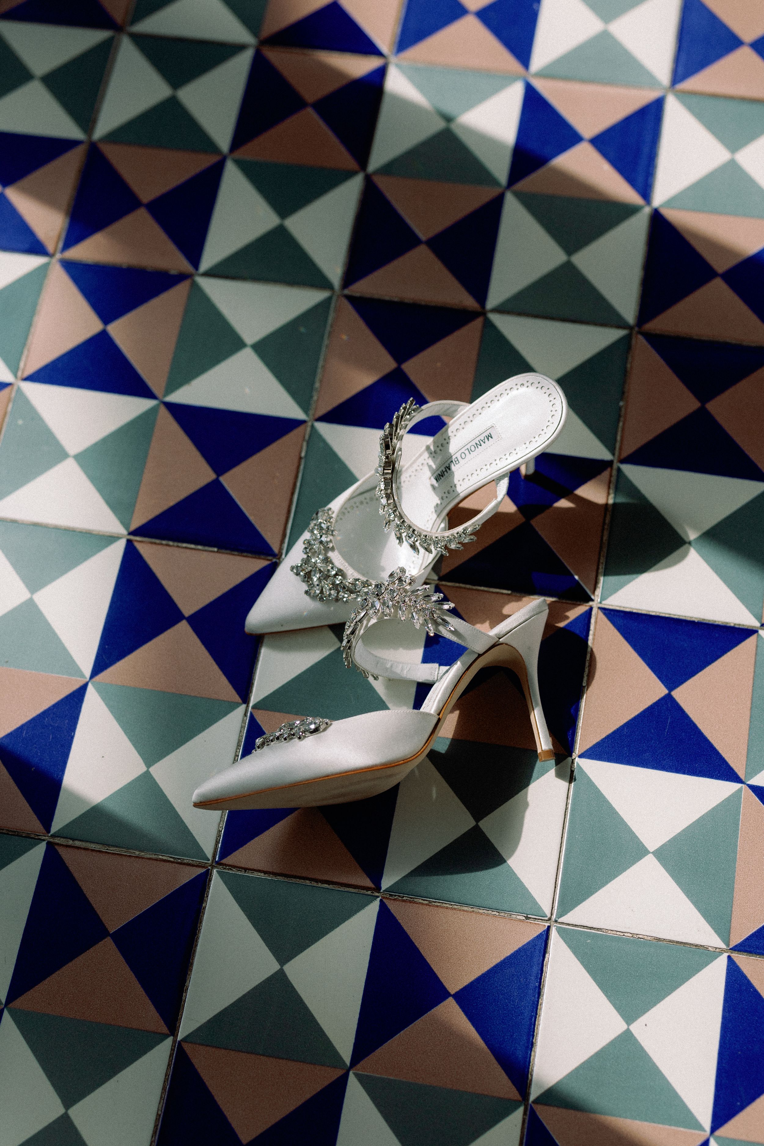 Elegant white bridal heels with crystal embellishments displayed on colorful geometric tile floor.