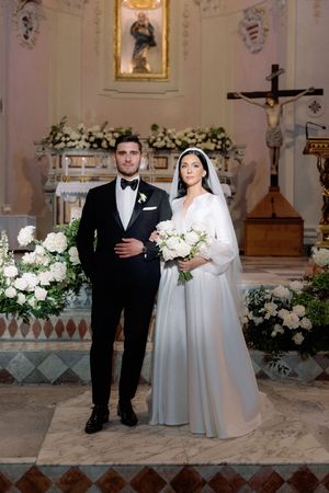 Bride and groom pose at church altar, she wears a white gown with veil, he wears a black tuxedo.