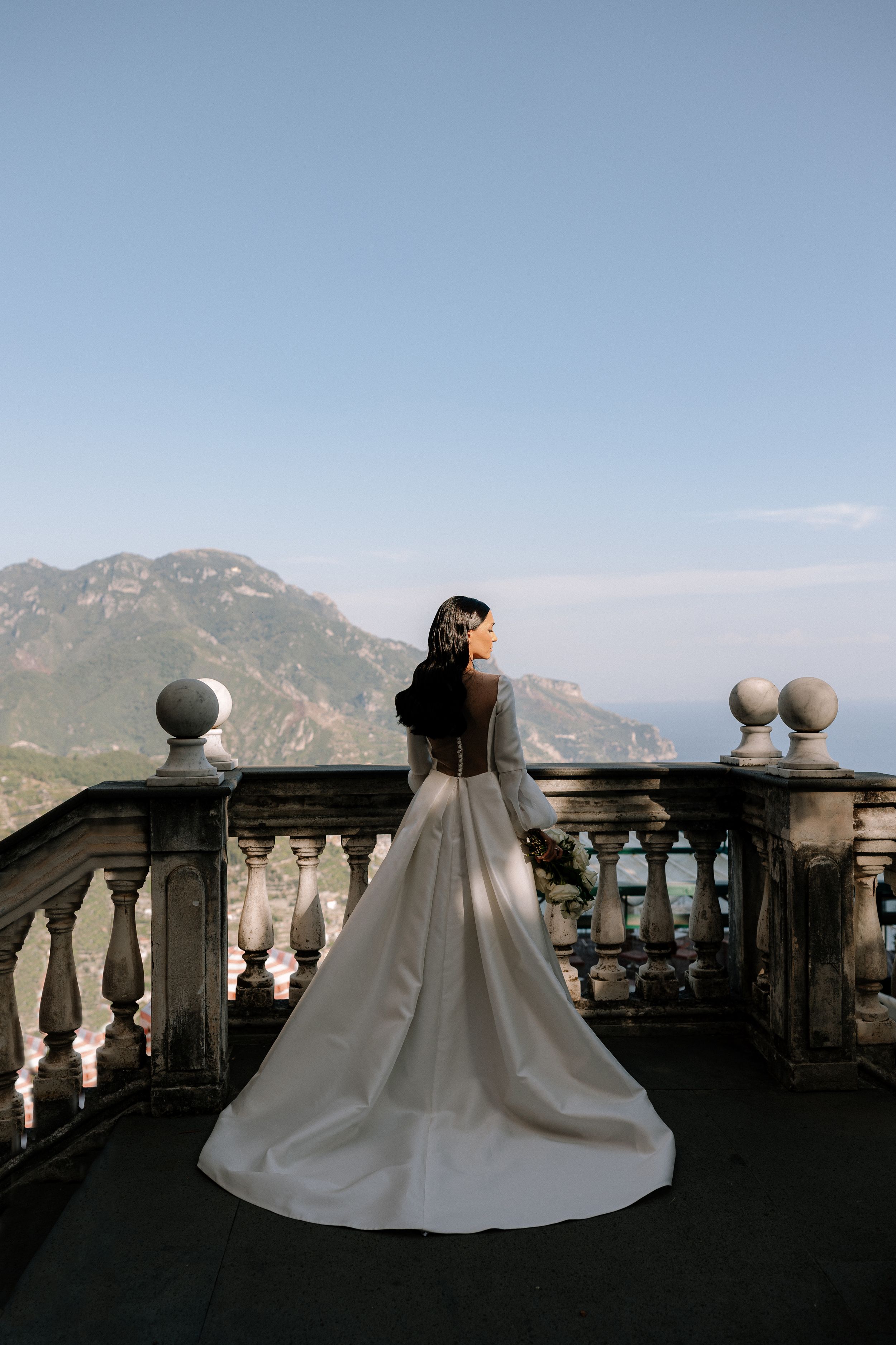 Bride in white gown with open back stands on stone balcony overlooking scenic mountain and sea views.