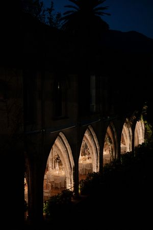 Gothic arched colonnade illuminated at night revealing an elegant wedding reception dining setup with warm glowing lights.