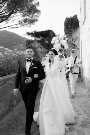 Black and white photo of a smiling bride and groom walking arm in arm on a scenic Italian coastal path.