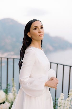 Elegant bride in white long-sleeve gown stands on balcony with mountain and coastal views behind her.