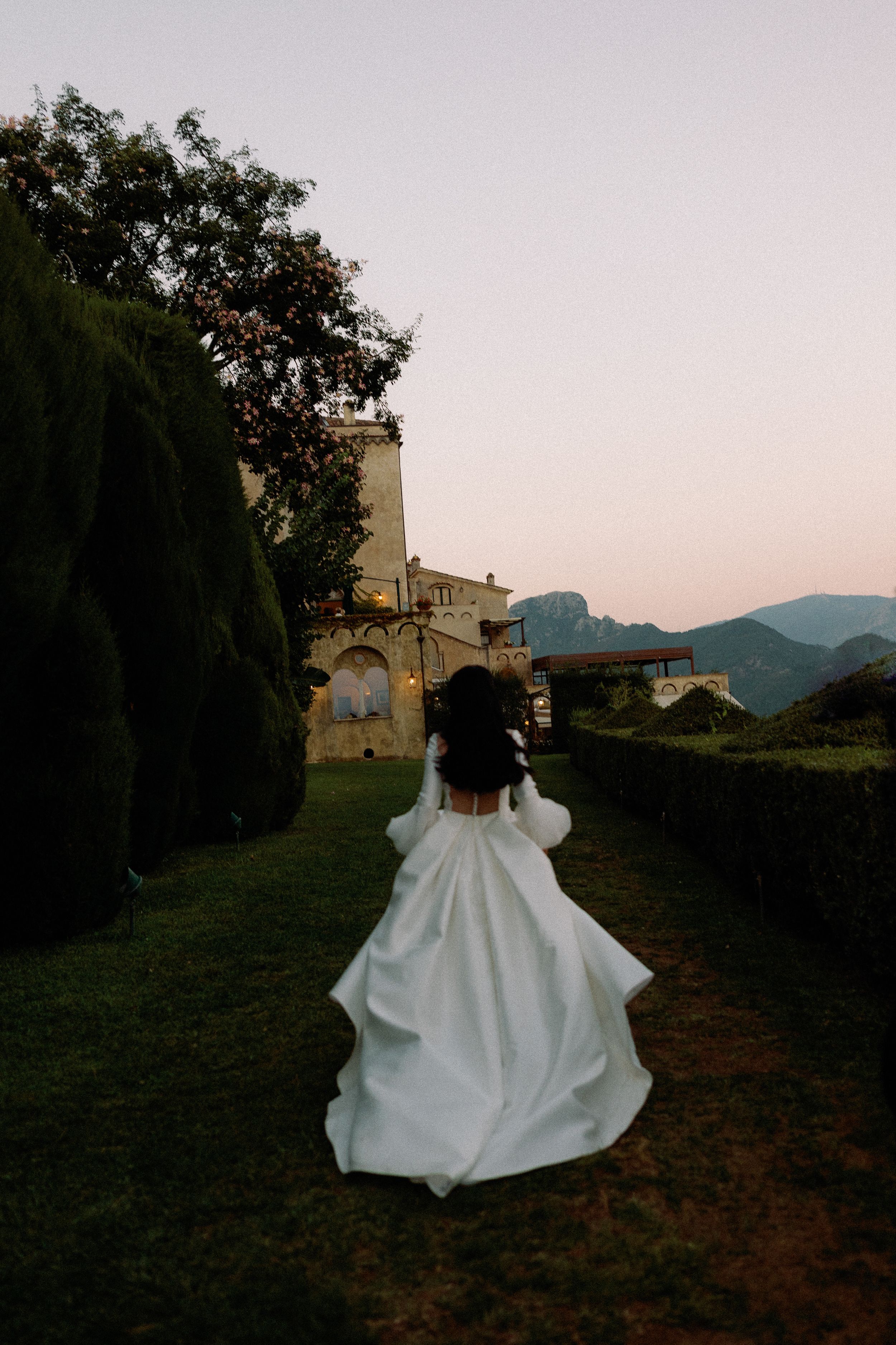 Bride in white gown with puff sleeves running through manicured garden toward historic castle at sunset.