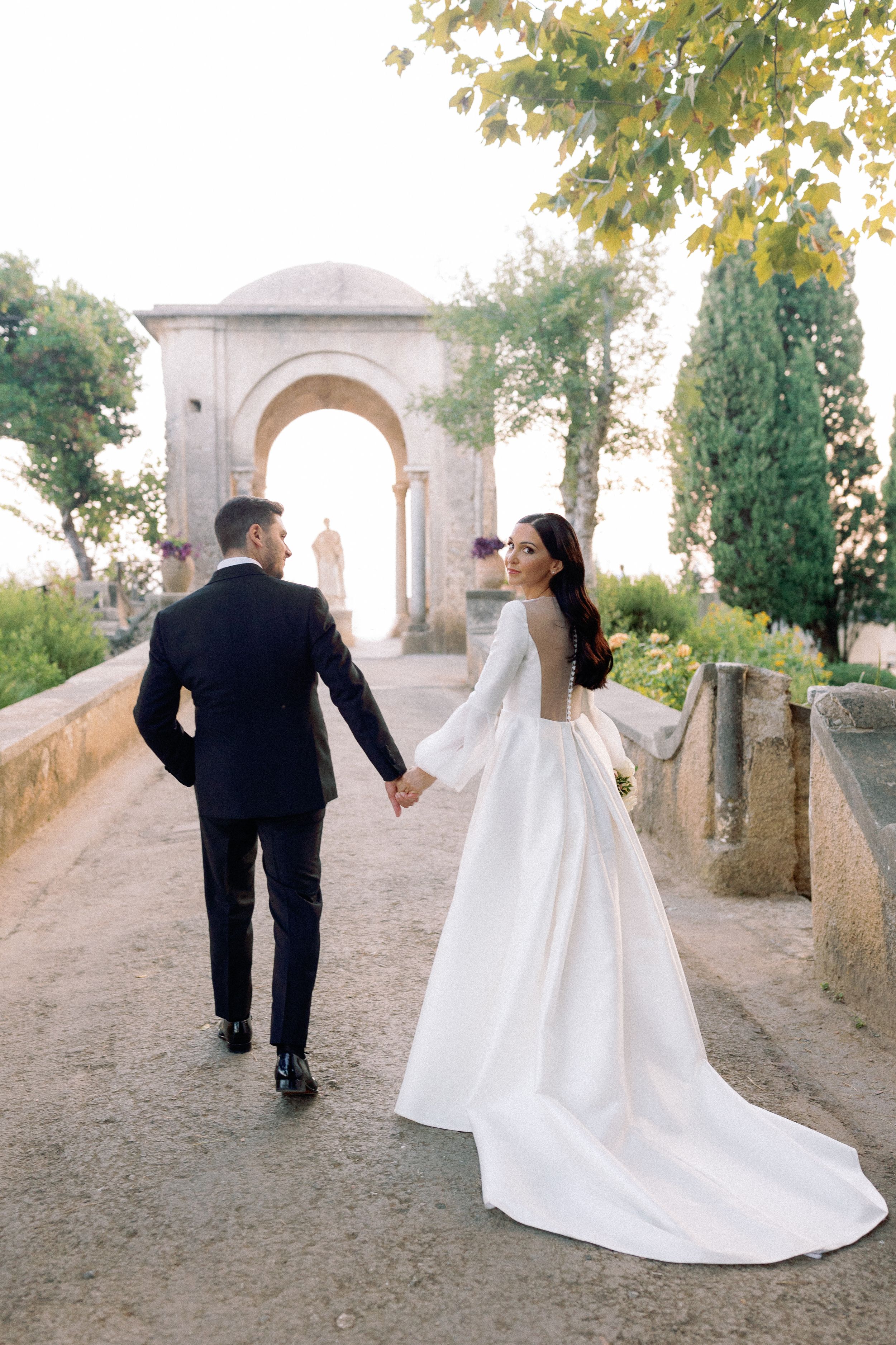 Bride and groom walk hand-in-hand toward a stone arch in a lush garden, bride wearing an elegant backless white gown.