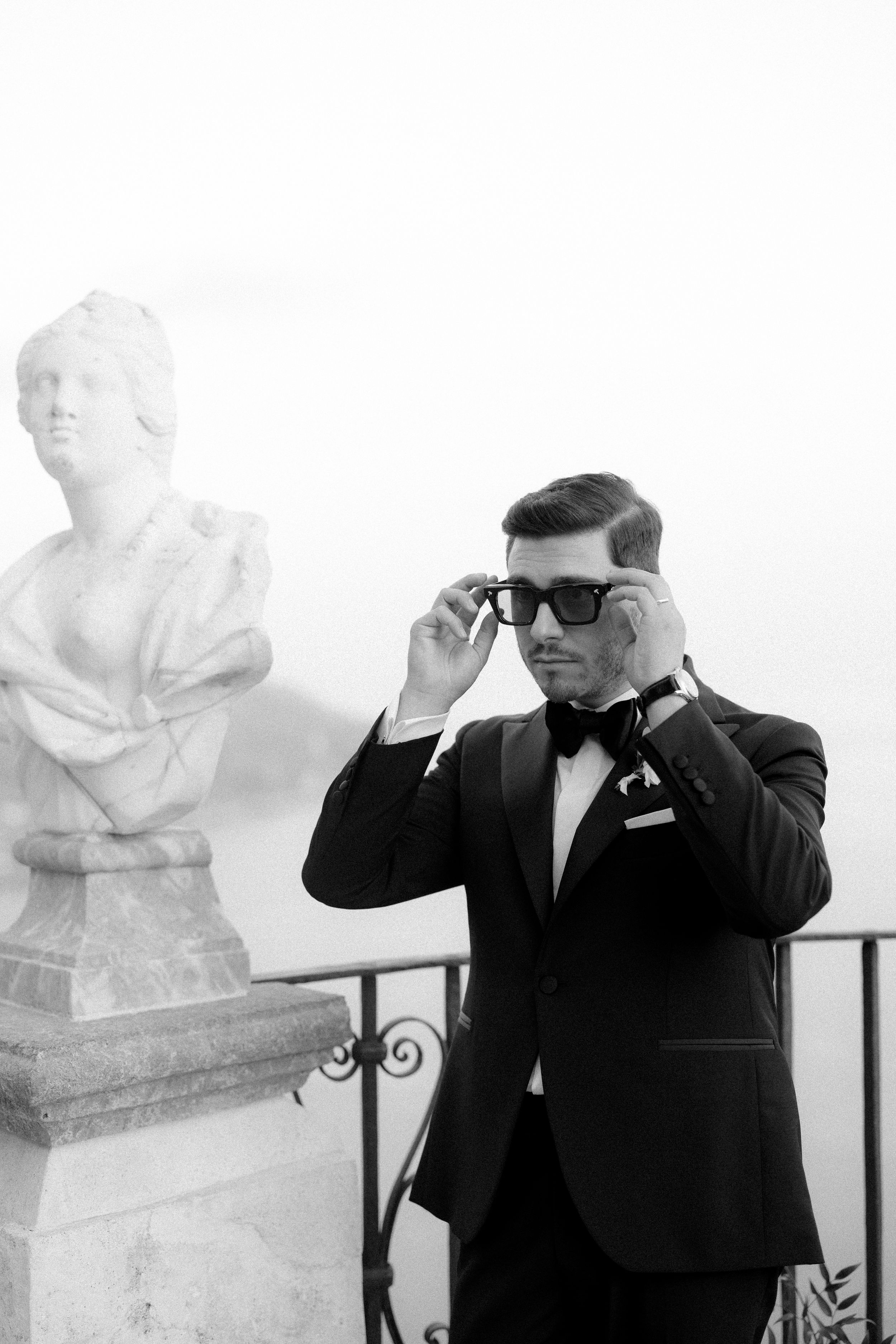 Elegant groom in tuxedo adjusting sunglasses beside classical marble bust sculpture in black and white wedding photo.