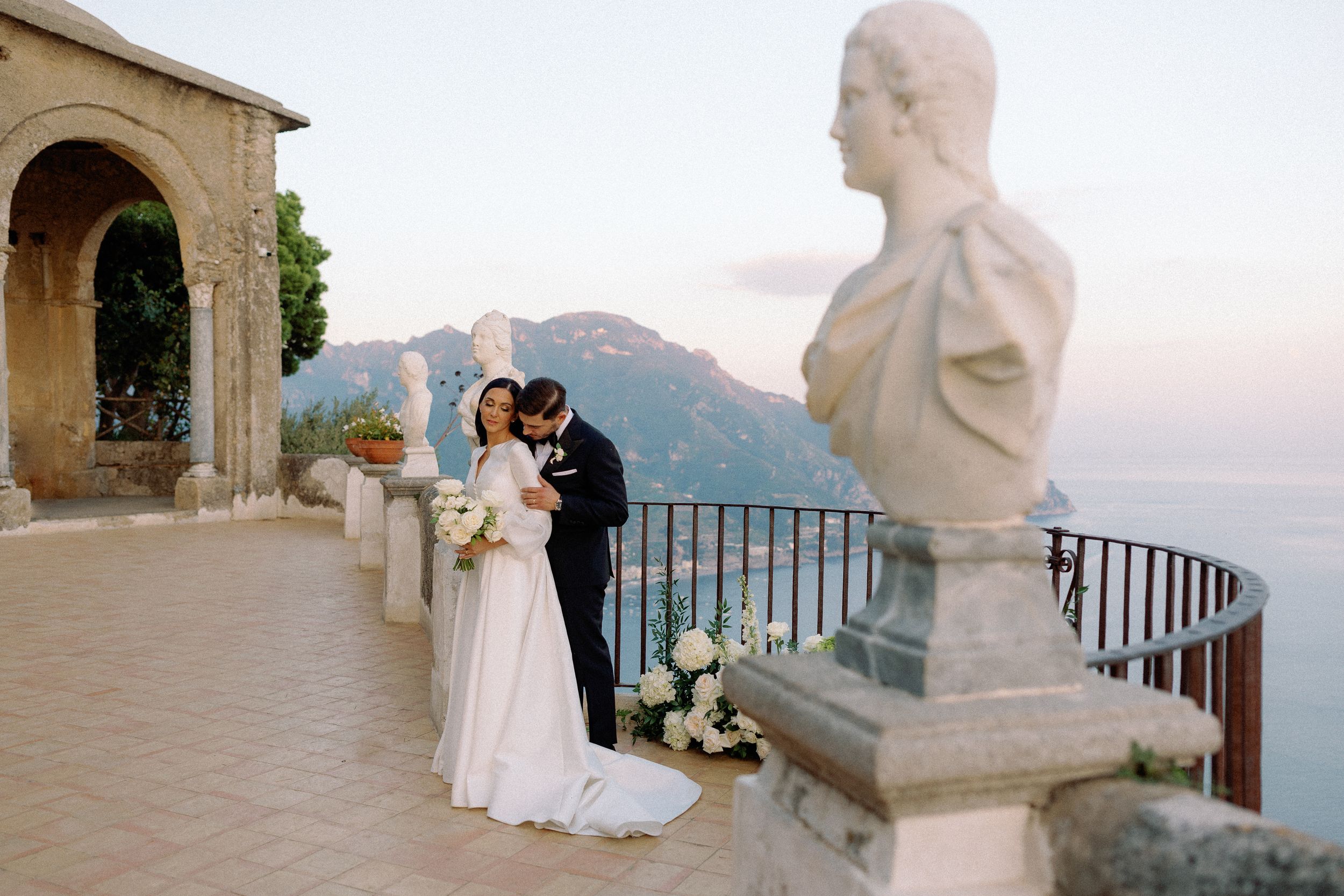 Bride and groom share a romantic moment on a terrace with marble busts and stunning Amalfi Coast views.