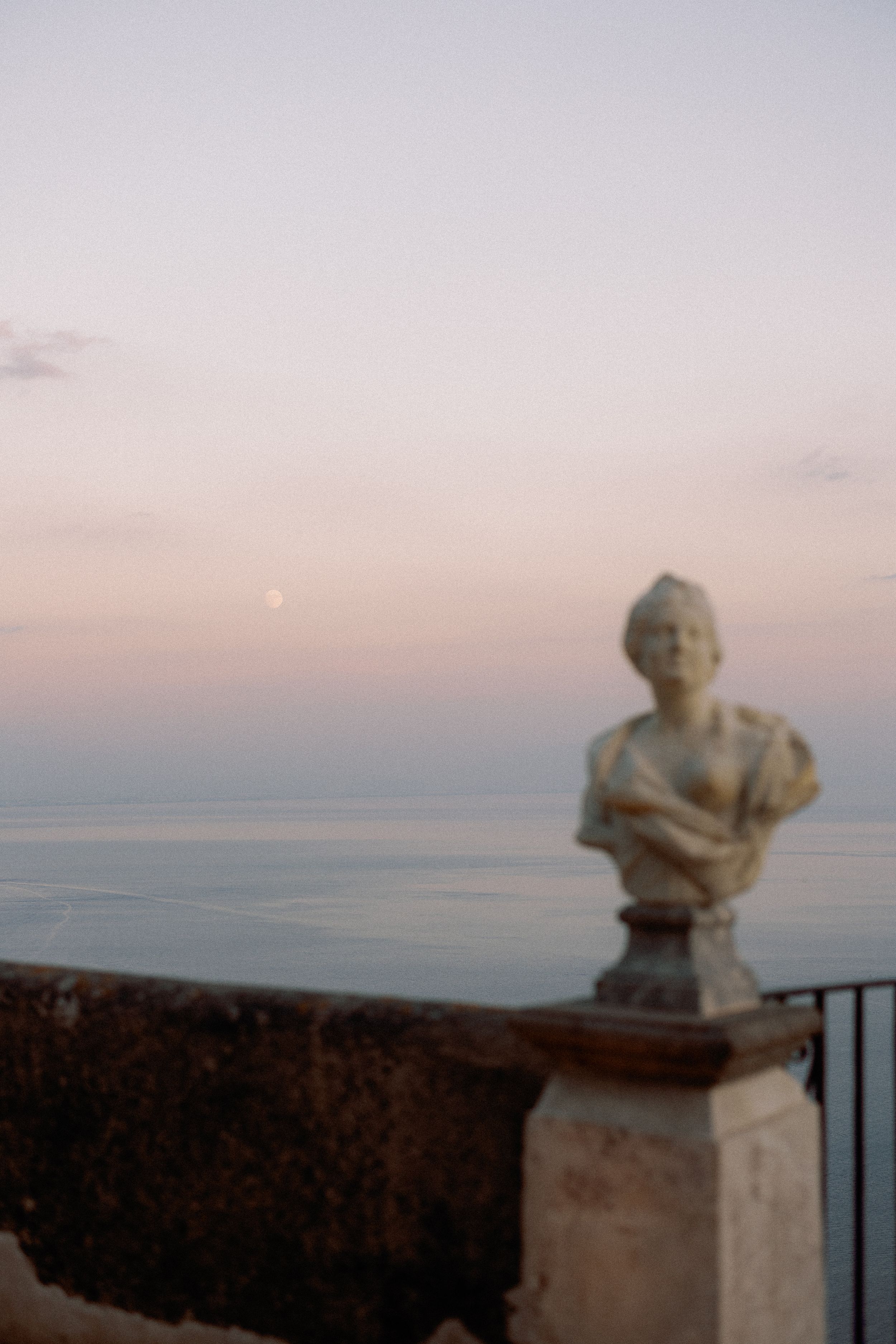 Classical bust statue on terrace overlooking calm sea at dusk with moon visible in pastel sky.