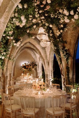 Elegant wedding reception in stone arched venue with crystal chandelier, floral arrangements, candlelit tables, and white chiavari chairs.