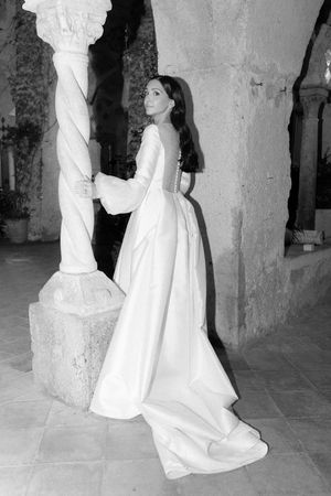 Bride in elegant long-sleeve white wedding gown posing beside ornate stone column in black and white photo.