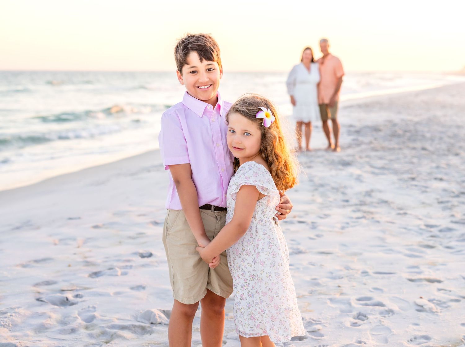 Two children in pastel outfits smile on a sandy beach at sunset with family in the background.