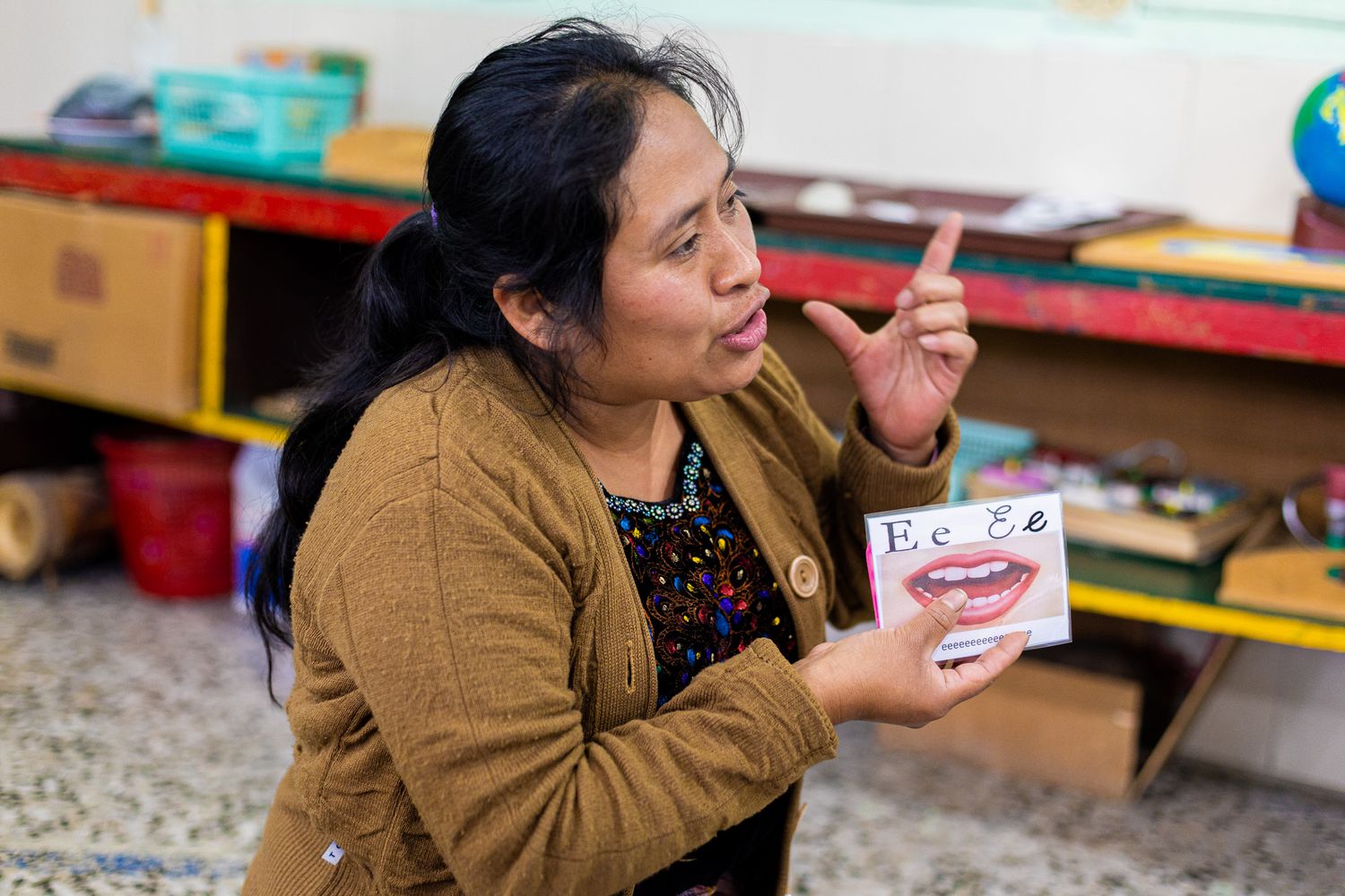 Teacher holding a phonics card showing letter E while pointing to her mouth demonstrating pronunciation in a classroom.