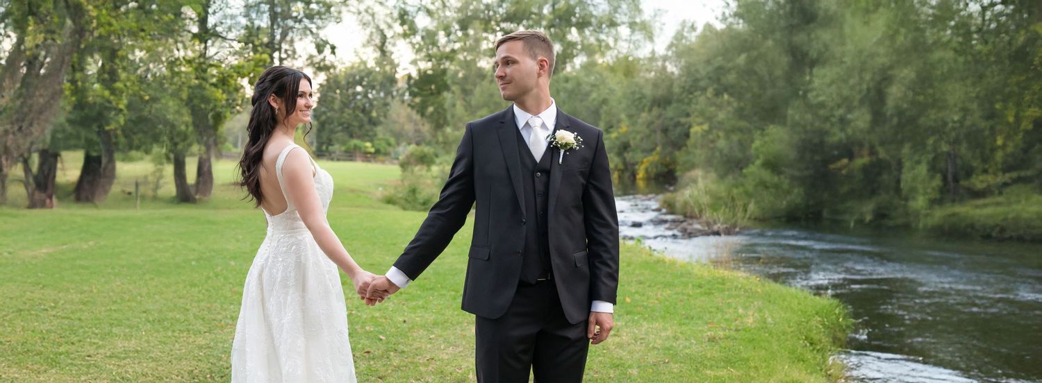 Bride and groom holding hands in lush green riverside garden setting.