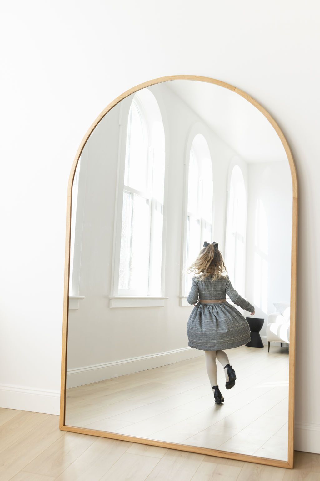 Young girl in plaid dress reflected in large arched gold-framed floor mirror leaning against white wall.