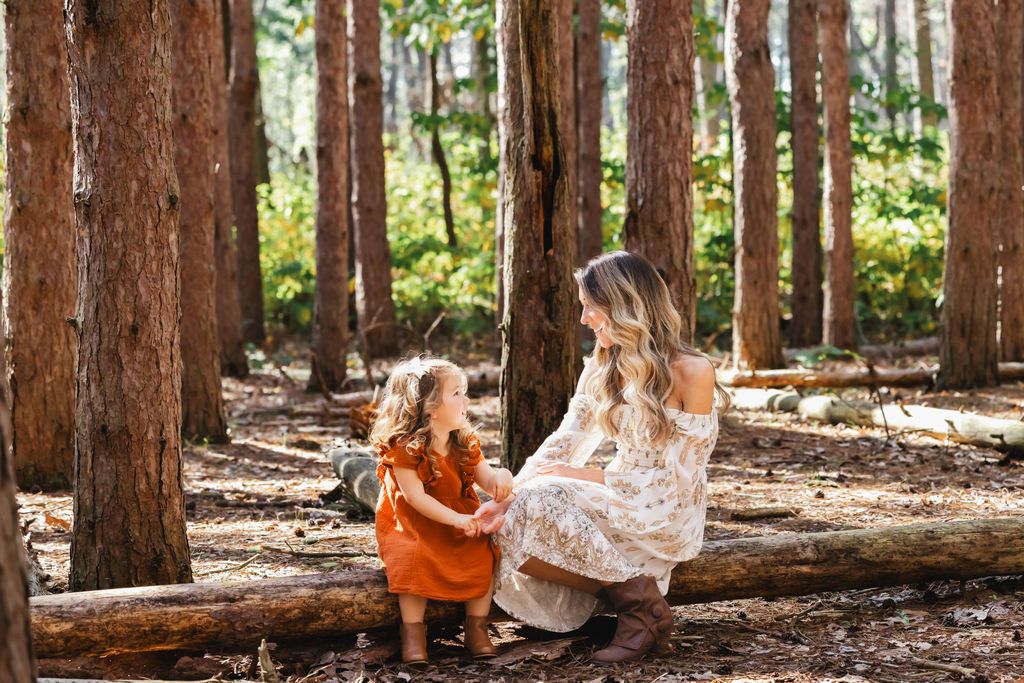 A mother and young daughter share a tender moment sitting on a fallen log in a sunlit redwood forest.