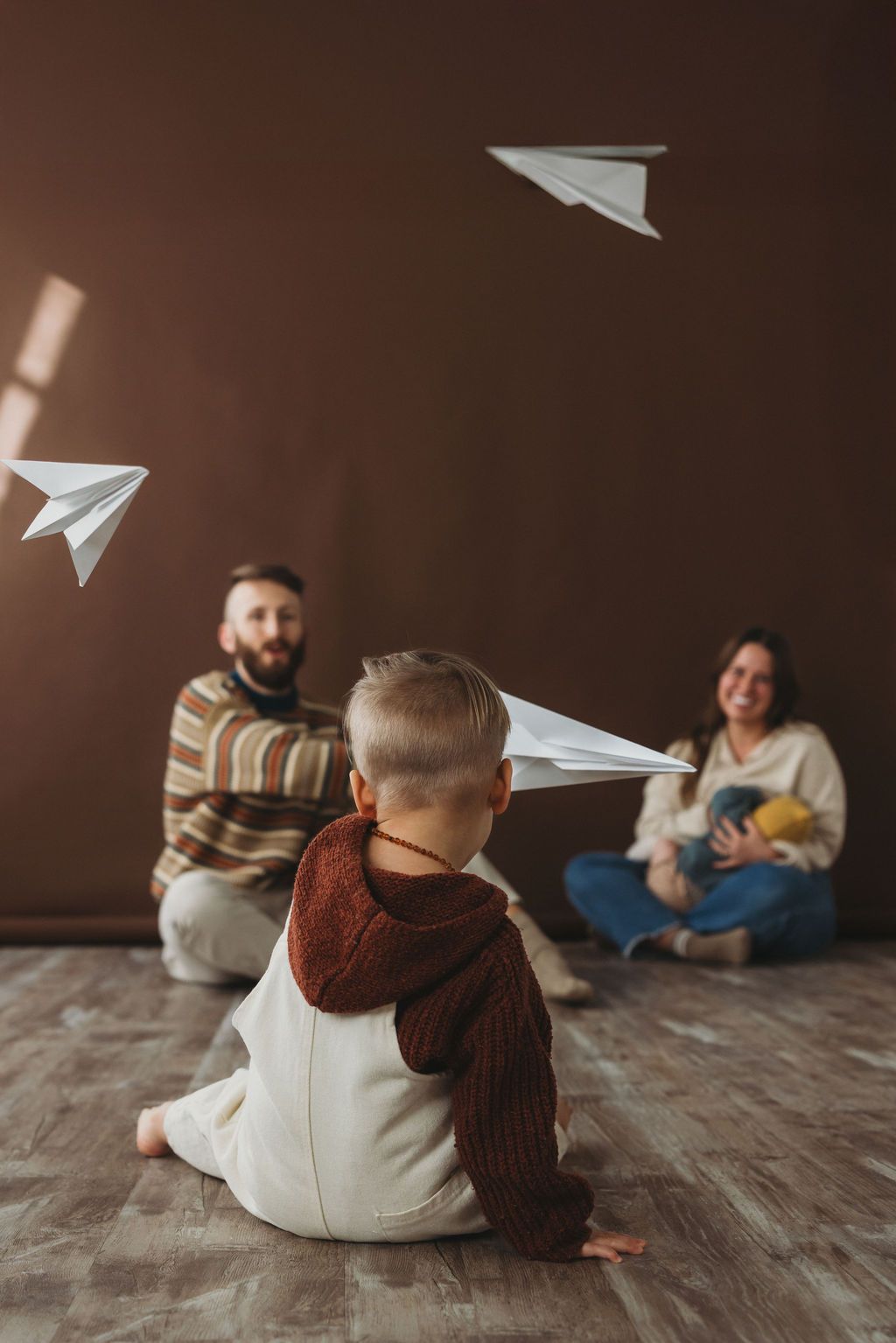 A joyful family sits on a wooden floor tossing paper airplanes with their toddler against a warm brown wall.