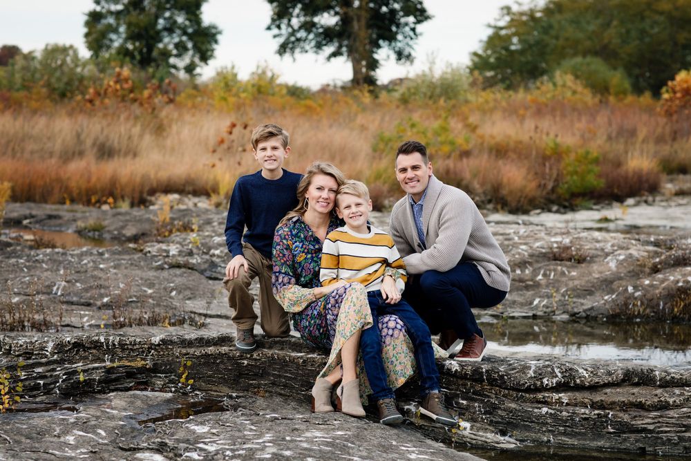 Family of four posing on rocks in an autumn field, smiling together during an outdoor fall family portrait session.