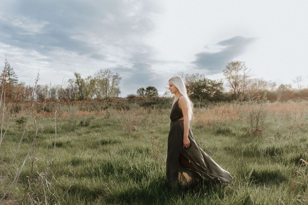 A blonde woman in a flowing dark maxi dress walks through a lush green meadow under a cloudy sky.