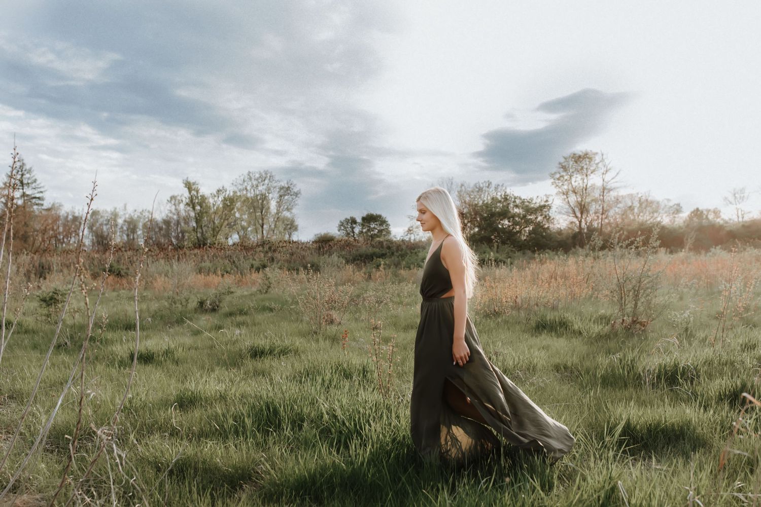 A blonde woman in a flowing dark maxi dress walks through a lush green meadow under a cloudy sky.