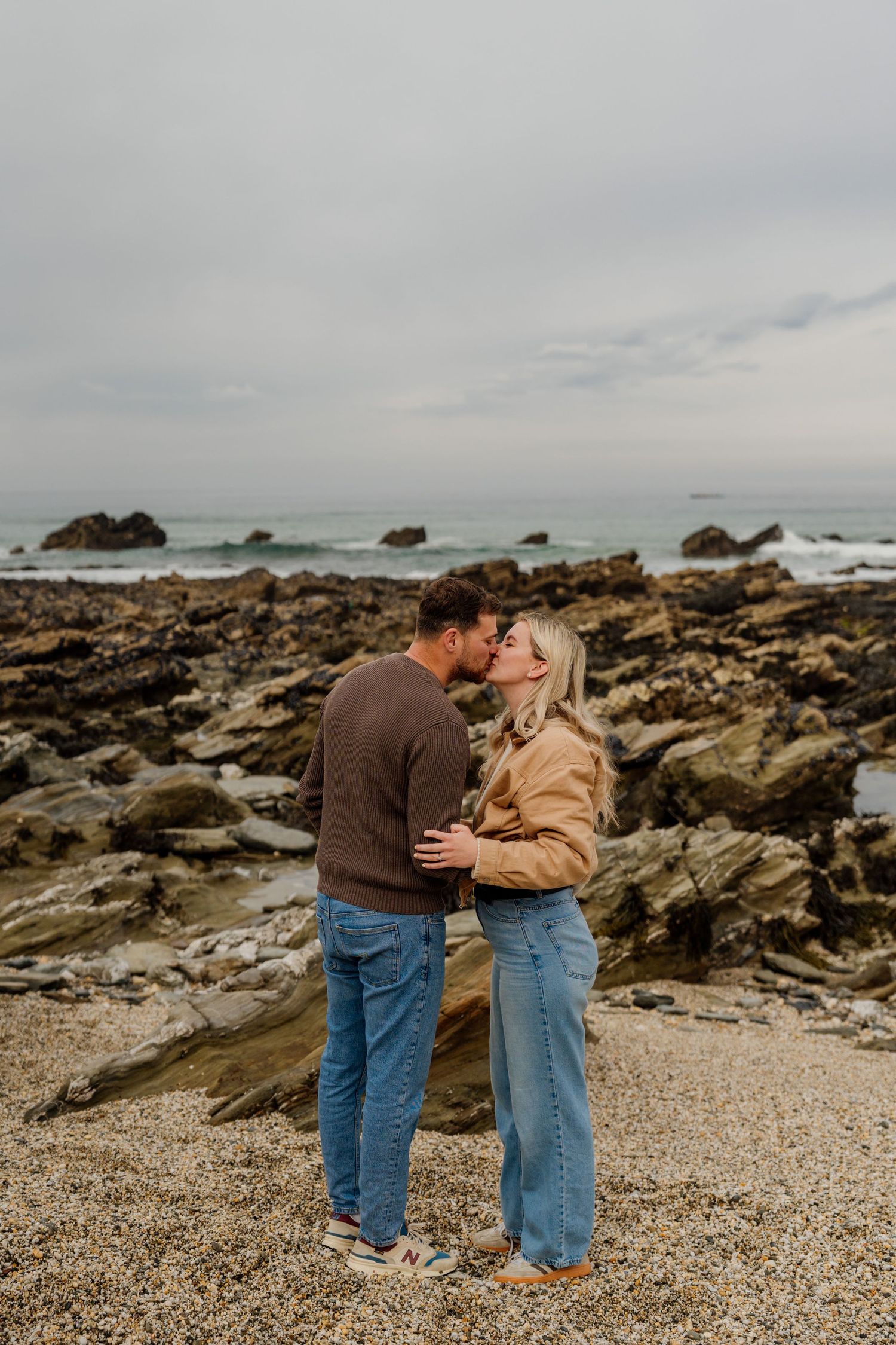 A couple shares a romantic kiss on a rocky beach with ocean waves and overcast skies behind them.