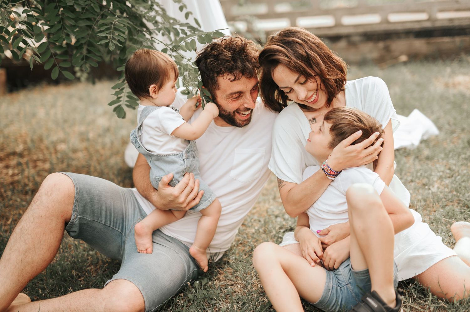 Happy young family with two toddlers sitting and laughing together on grass in outdoor setting.