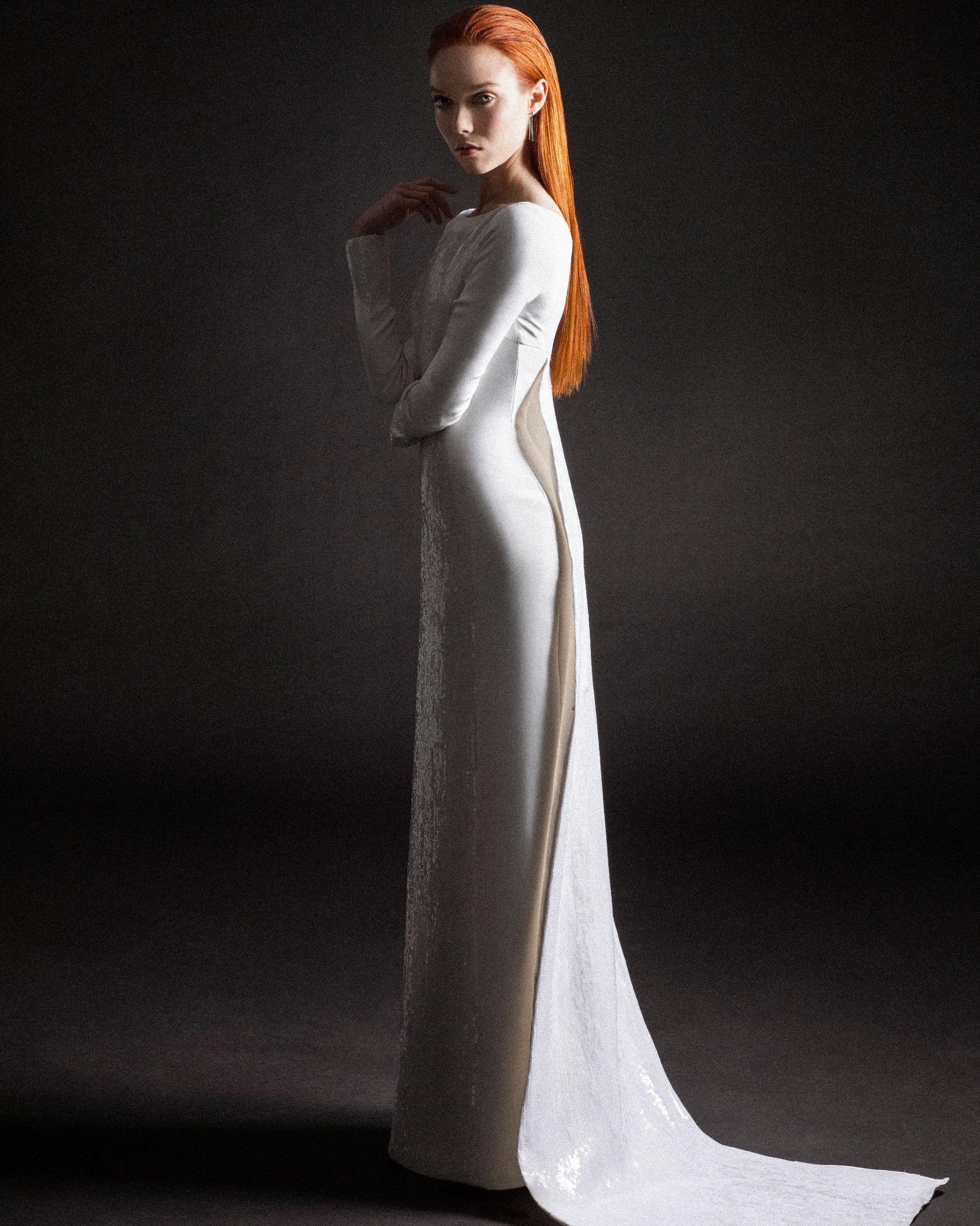 A woman in a white long-sleeve gown with a train poses gracefully, her long red hair flowing over her shoulder.