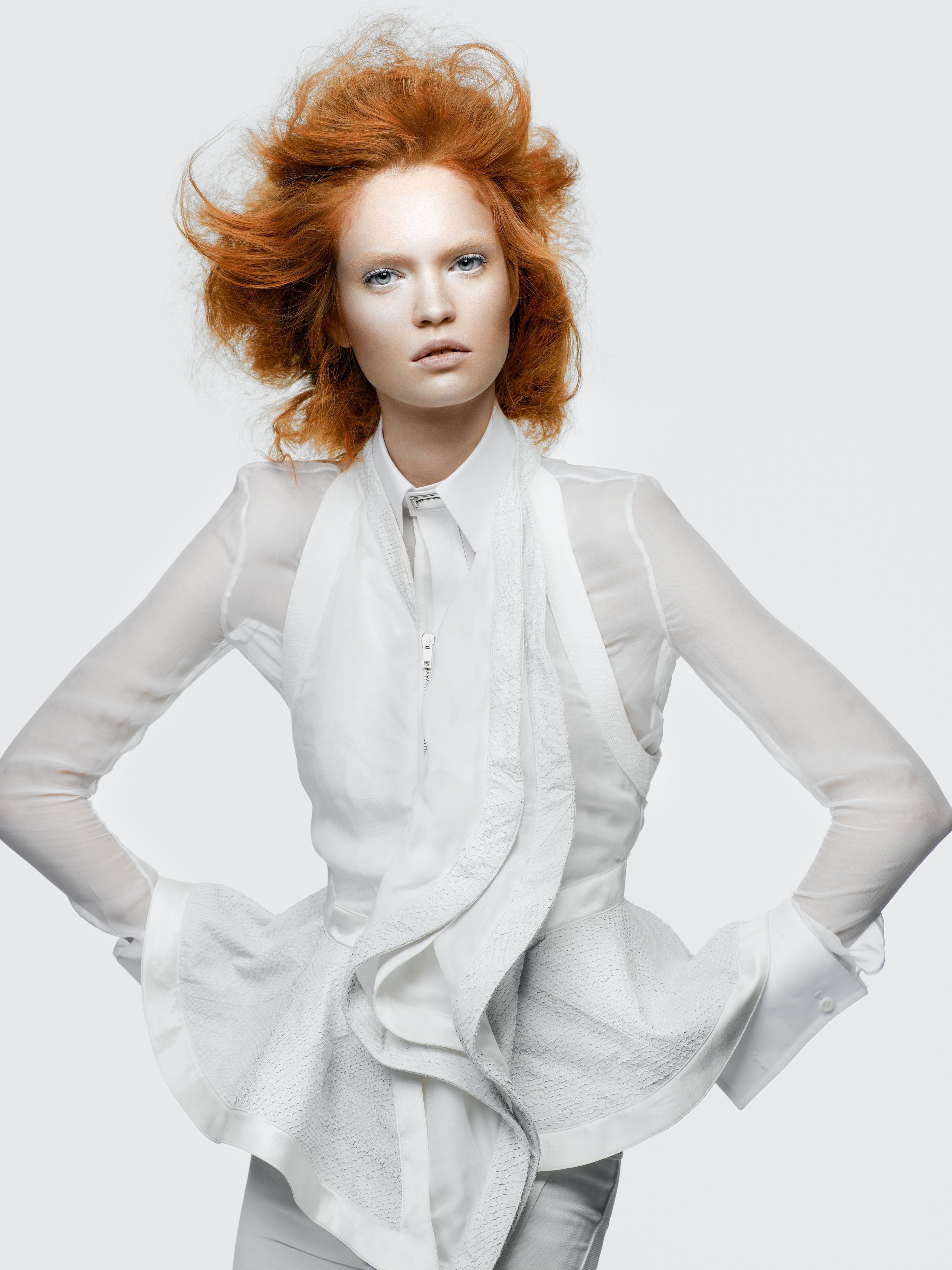 A striking female model with voluminous red hair poses in an all-white layered outfit against a light background.