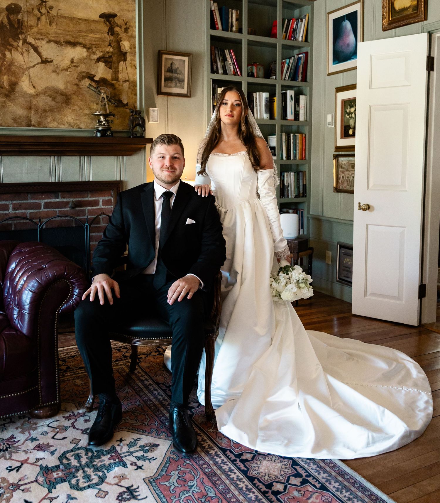 Elegant wedding portrait of bride in white gown with train and groom in black tuxedo posing in a classic library setting.