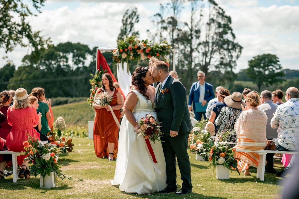 Bride and groom share first kiss at outdoor wedding ceremony with floral arch and seated guests.