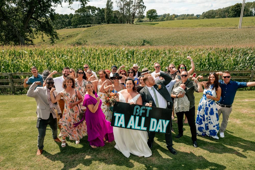 Wedding group photo on lawn holding BFT TA ALL DAY banner with cornfield backdrop on sunny day.