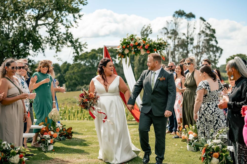 Bride and groom walk down outdoor aisle at rustic farm wedding, guests cheering, floral arch backdrop.
