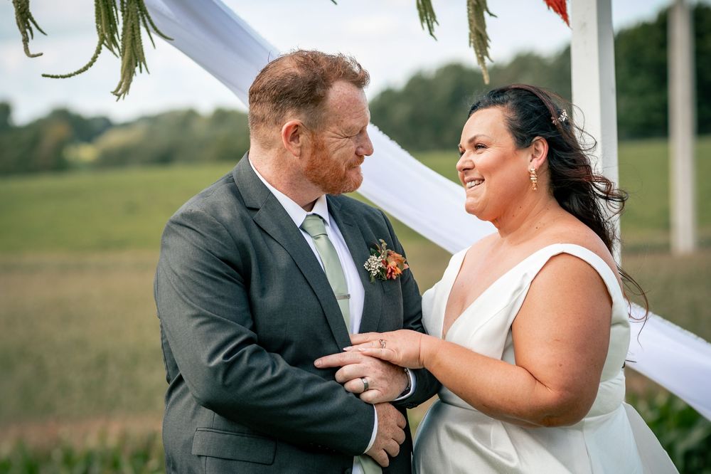 Couple exchanging vows at outdoor wedding ceremony, bride in white gown holding hands with groom in gray suit.