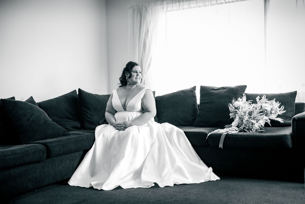 Bride in a white gown sits elegantly on a dark sofa with a bouquet beside her in black and white.