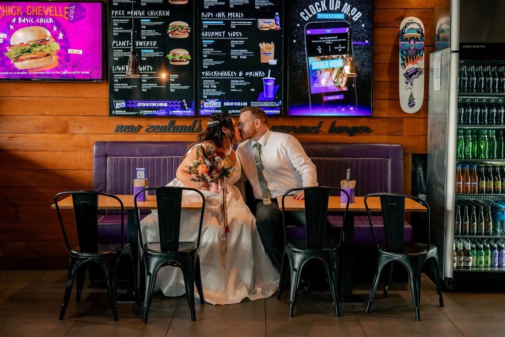 Bride and groom share a kiss at a burger bar restaurant, surrounded by neon signs and industrial decor.