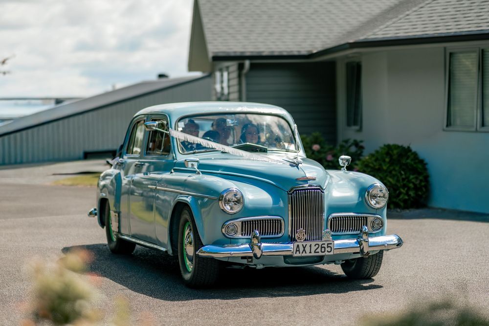 Vintage blue classic car decorated with white ribbons parked outside a building on a sunny wedding day.