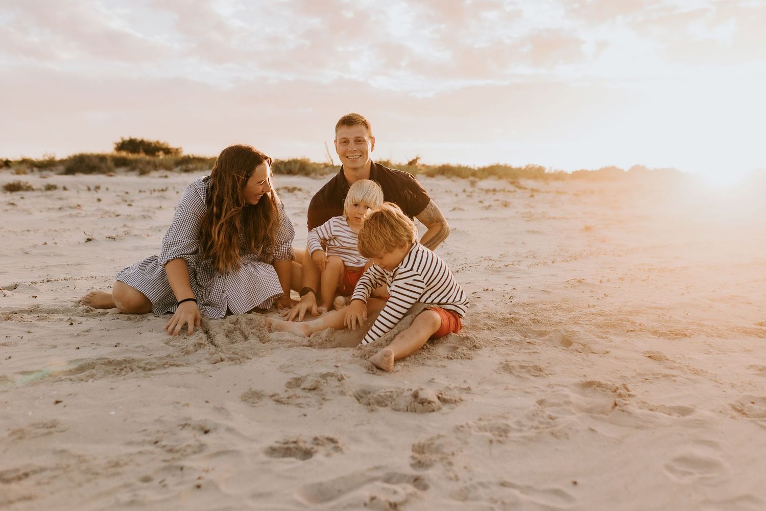 Family sitting together on sandy beach at golden sunset, two adults and two young children playing in the sand.