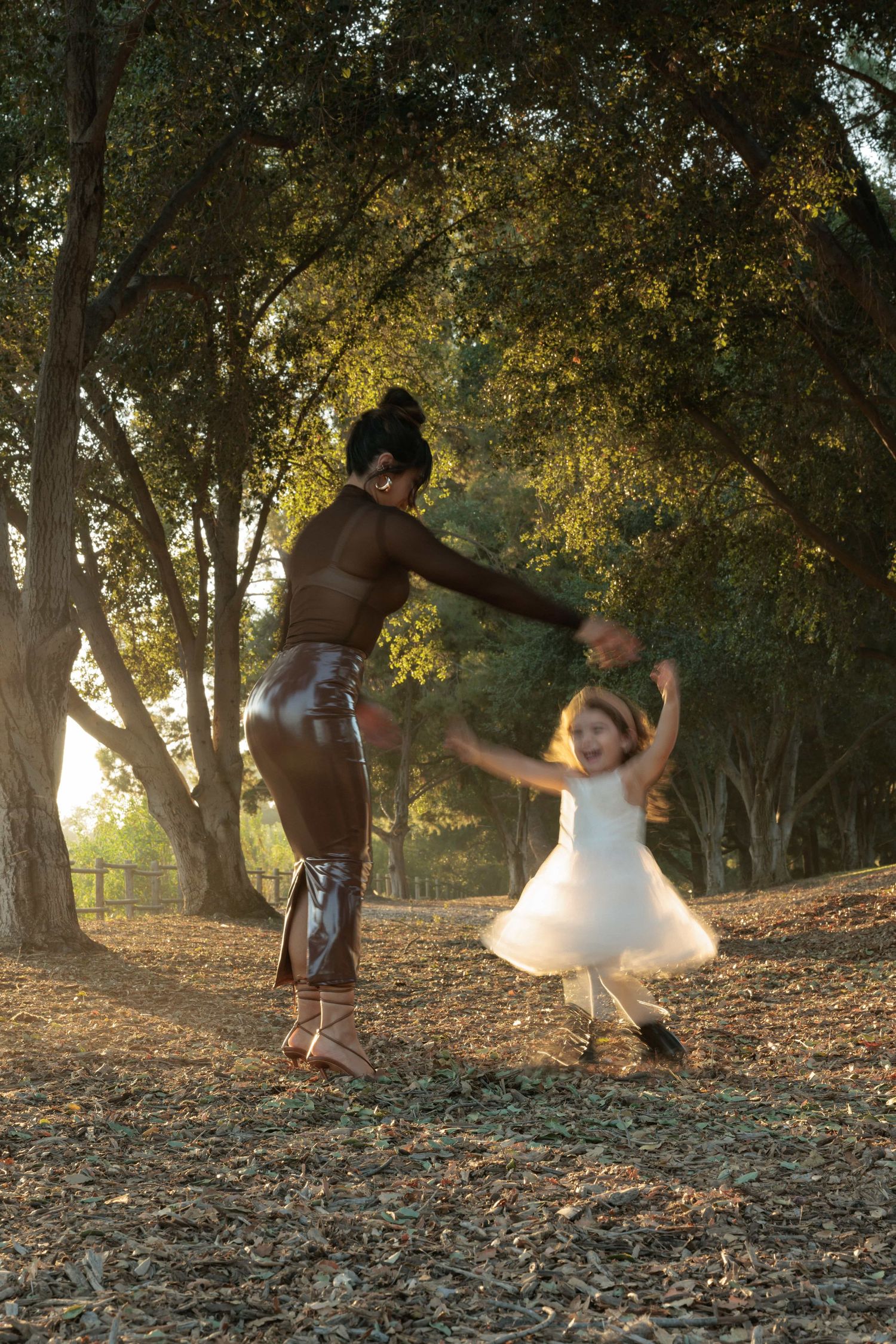 Woman and young girl in white dress dancing together in sunlit park surrounded by trees.
