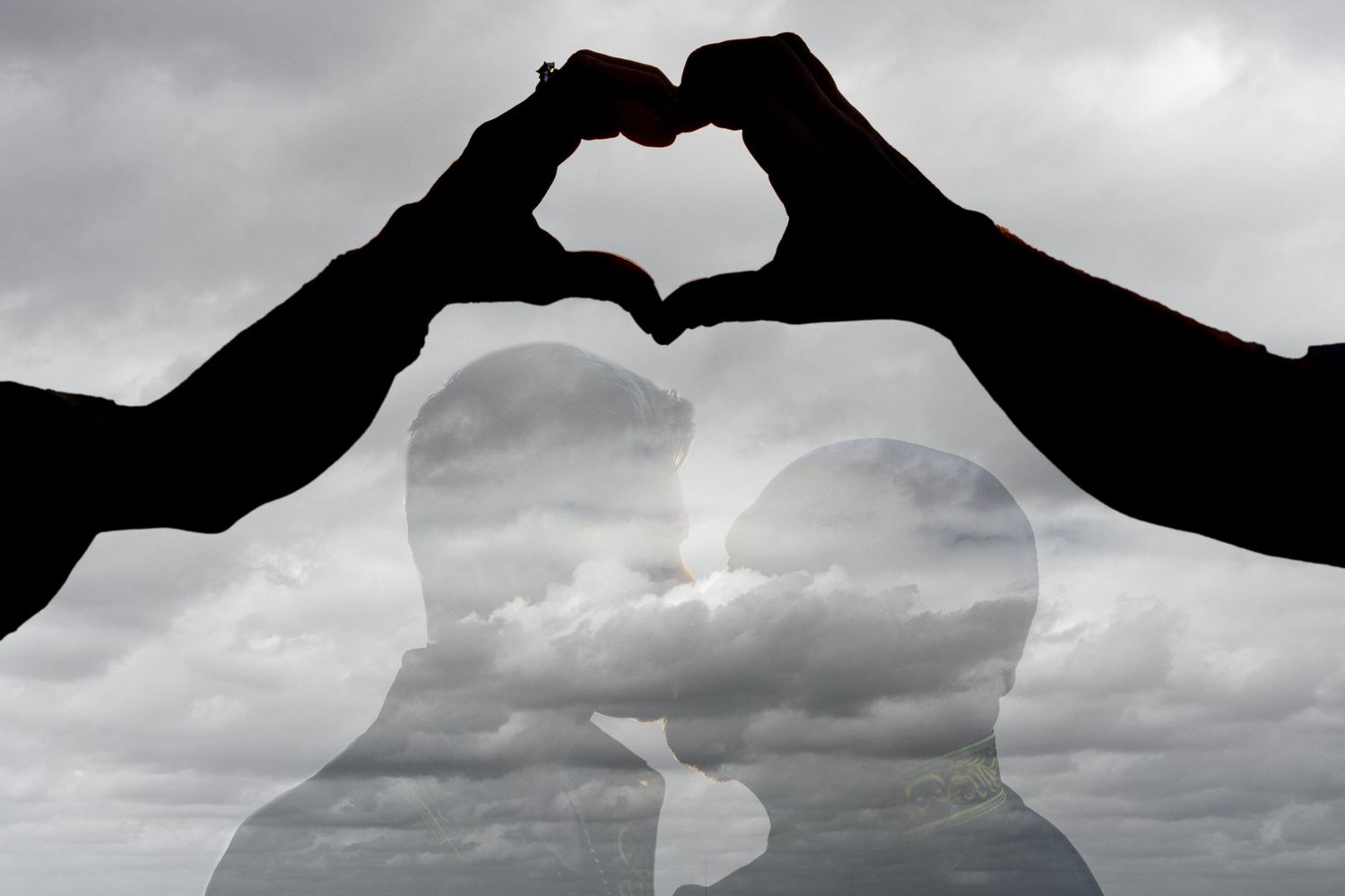Silhouette of couple kissing under heart-shaped hands against dramatic cloudy sky in romantic double exposure photo.