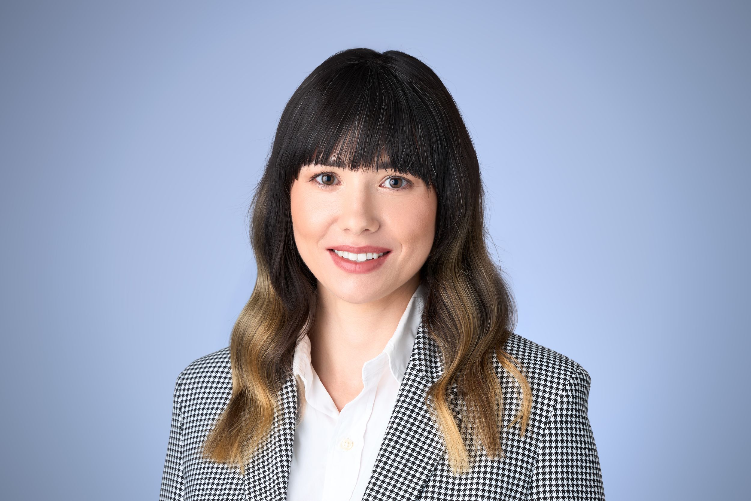 Professional headshot of a smiling woman with dark hair wearing a plaid blazer against a blue background.